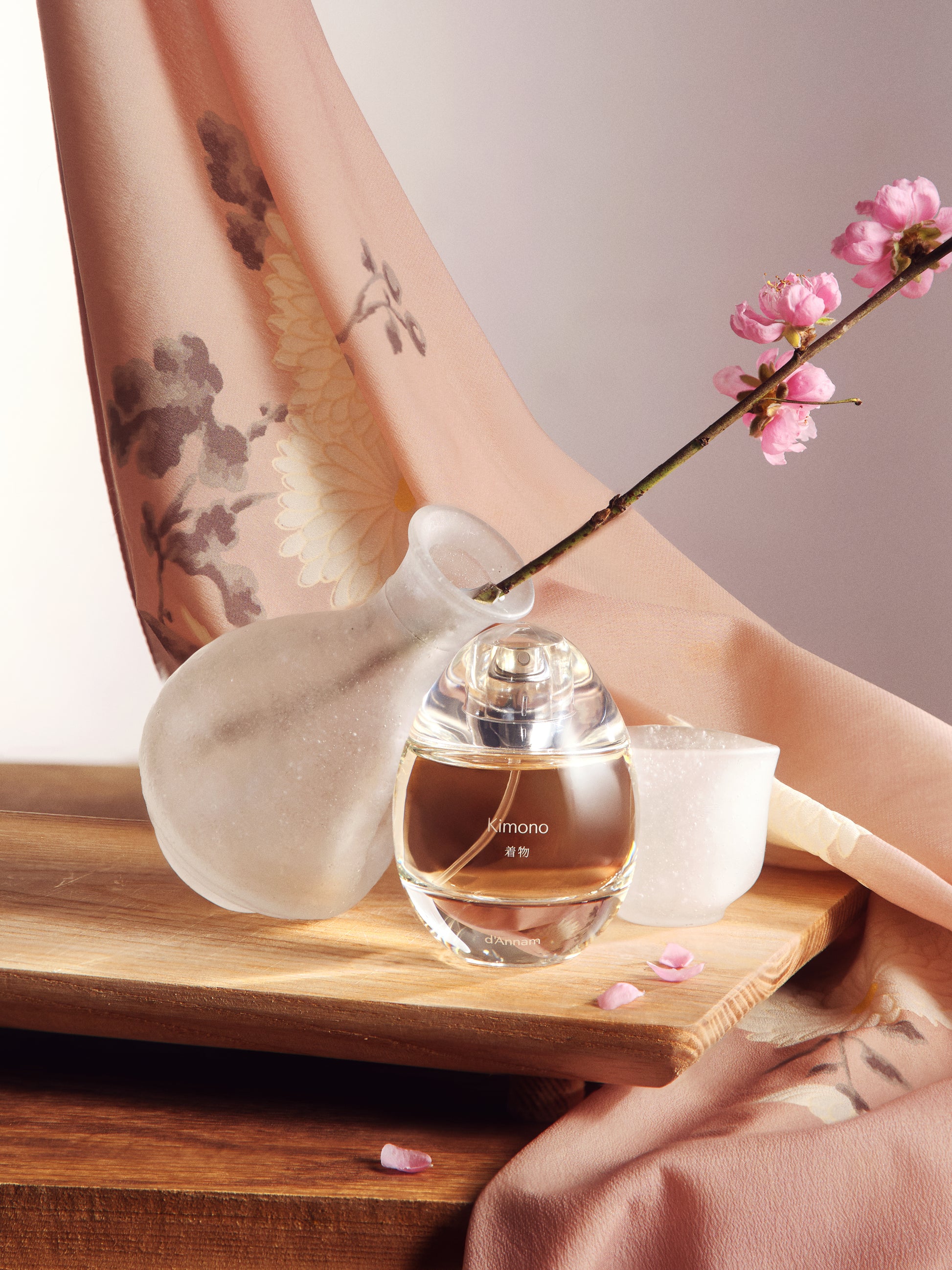 Perfume bottle on a wooden tray with a decorative vase and pink flowers, against a neutral background.
