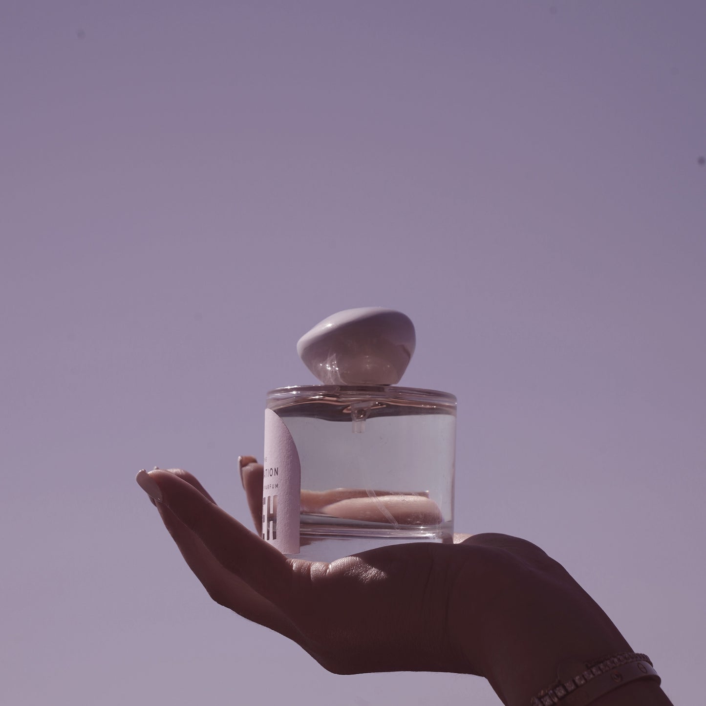 Hand holding a clear glass bottle with a stone on top against a purple sky
