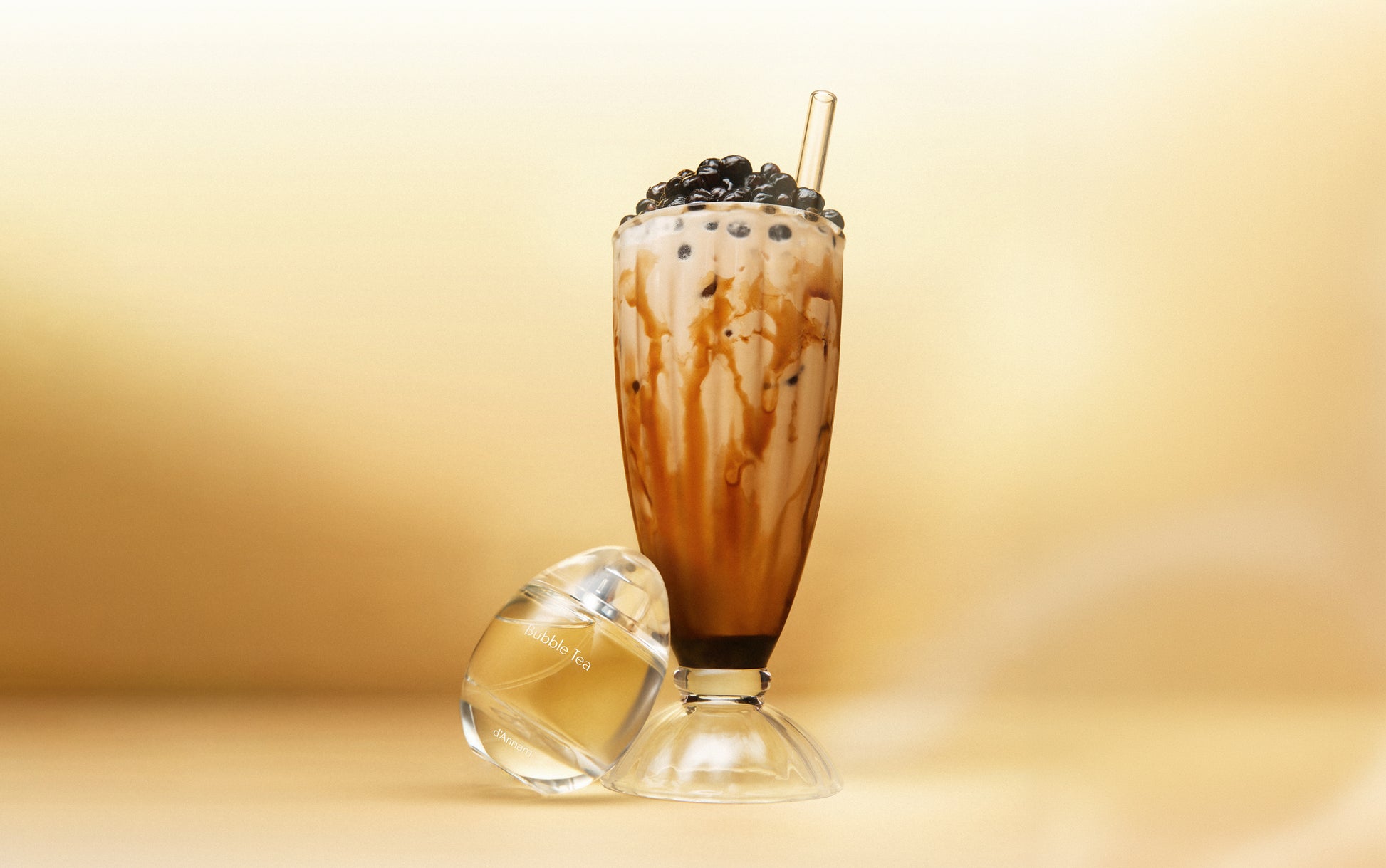 A tall clear glass filled with a milk-based drink topped with dark tapioca pearls and a straw, set against a warm beige background with a small clear glass container in front.
